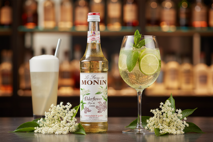 Monin Elderflower syrup bottle with a cocktail and a glass of elderflower drink on a bar counter.